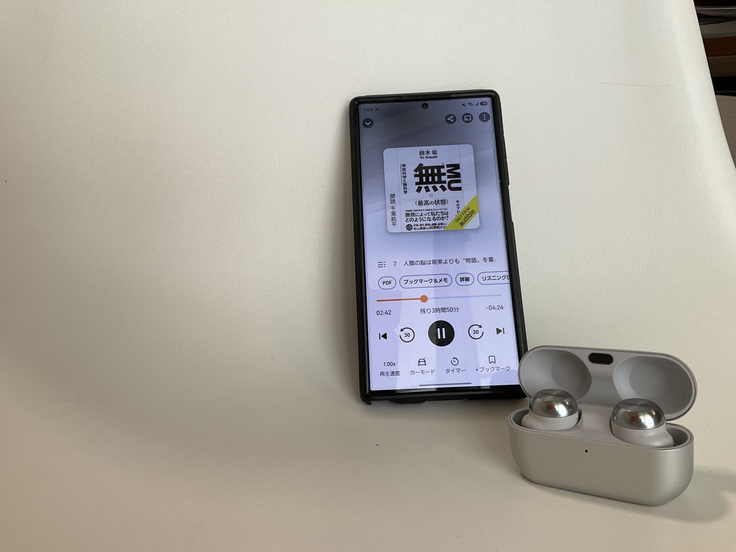 スマートフォンのAudibleアプリで再生中の画面と、横に置かれたワイヤレスイヤホンのケース